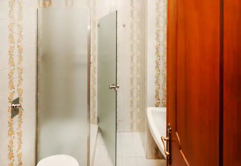 Baño pequeño con azulejos decorados, puerta de ducha de vidrio esmerilado y puerta de madera; distribución compacta y detalles vintage.
