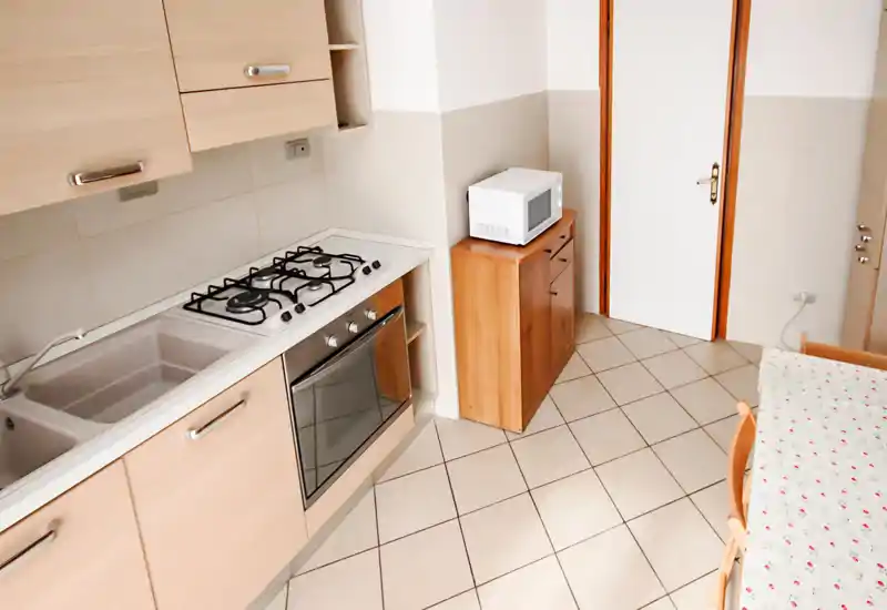 Cocina compacta y práctica con placa de gas, horno empotrado, fregadero doble y microondas sobre un mueble de madera. Baldosas neutras y muebles claros crean un área de cocina luminosa y funcional.