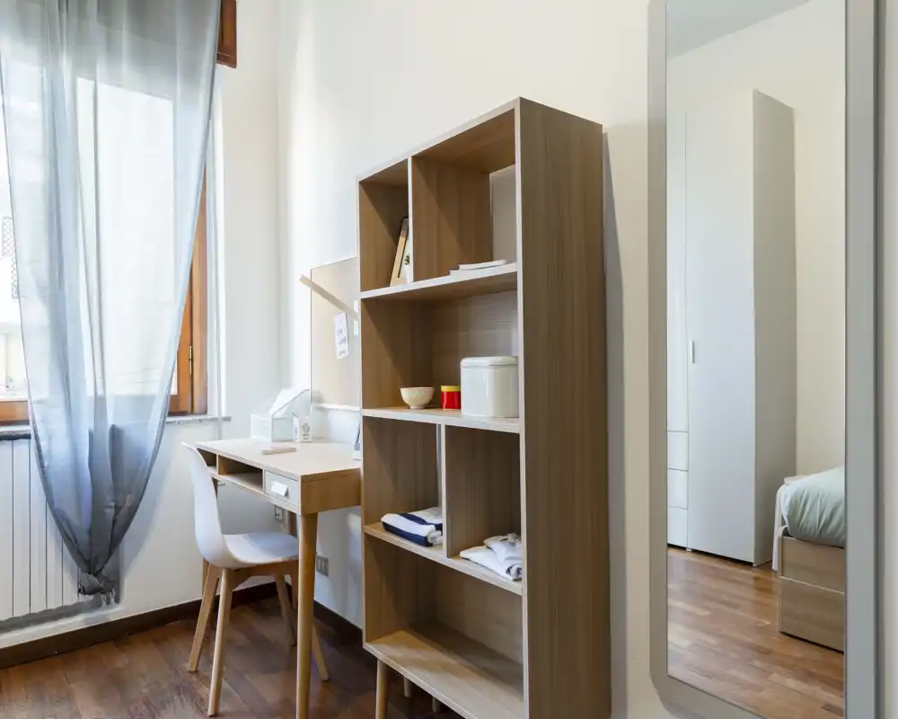 Rincón de dormitorio luminoso y minimalista con estantería de madera, pequeño escritorio junto a la ventana y espejo que refleja parte de la cama—limpio, moderno y bien iluminado.