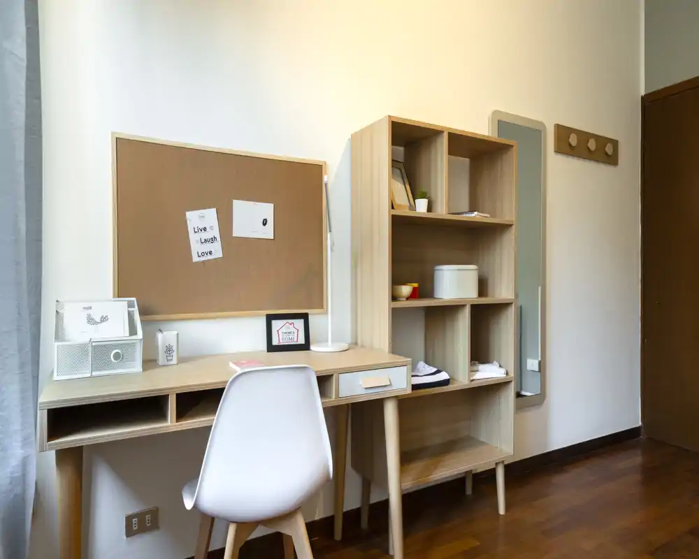 Zona de estar luminosa con escritorio compacto y estantería de madera, ideal como rincón de oficina en casa; las tonalidades cálidas y el corcho dan un toque personal.