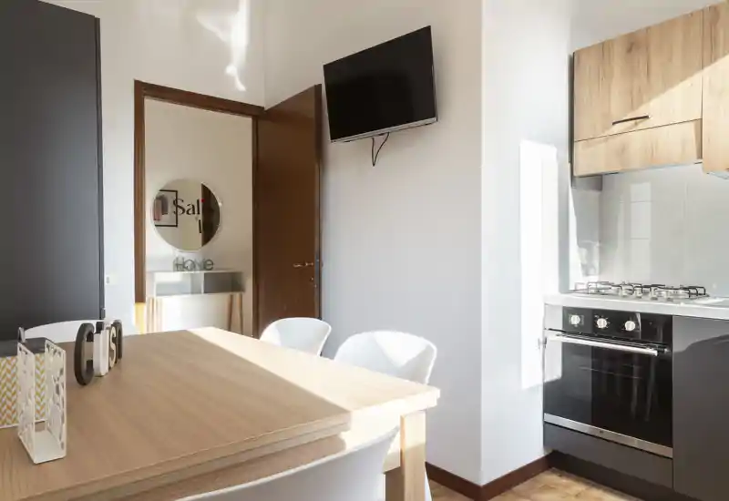 Angolo cucina moderno e luminoso con tavolo in legno, quattro sedie bianche e piano cottura a gas; luce naturale valorizza i pensili in legno e la TV a parete.