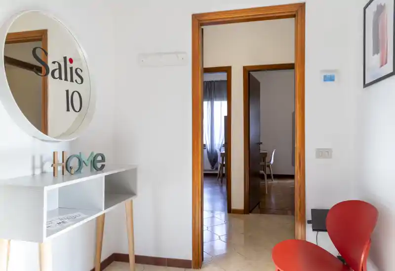 Piccolo ingresso luminoso con consolle, specchio tondo e una sedia rossa; cornici in legno aprono su stanze adiacenti.