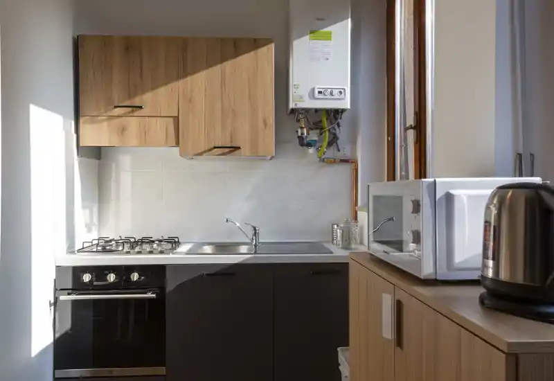 Cucina moderna compatta con mobili in legno, piano cottura a gas, lavello e piccoli elettrodomestici sul piano. La luce naturale da una finestra alta illumina bene l'ambiente, perfetta per un appartamento.