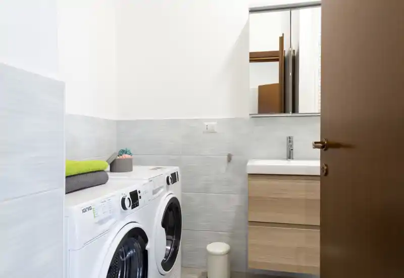 Bagno compatto con lavanderia integrata: lavatrice e asciatrice a carica frontale accanto al lavabo con mobile in legno e pensile con specchio. Spazio luminoso, pulito e funzionale, ideale per annunci di appartamenti.