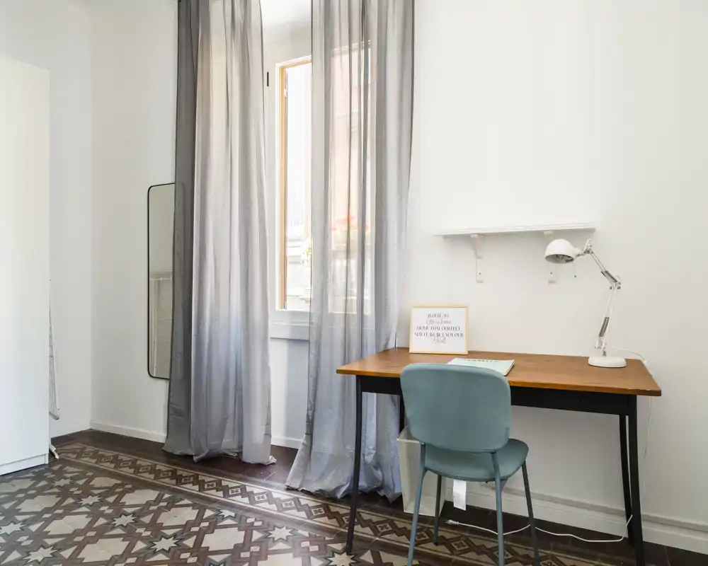 Rincón de habitación luminoso y minimalista con escritorio de madera, silla verde azulado y cortinas traslúcidas largas junto a la ventana; suelo de baldosas limpio y una lámpara sencilla crean un espacio de trabajo sereno.