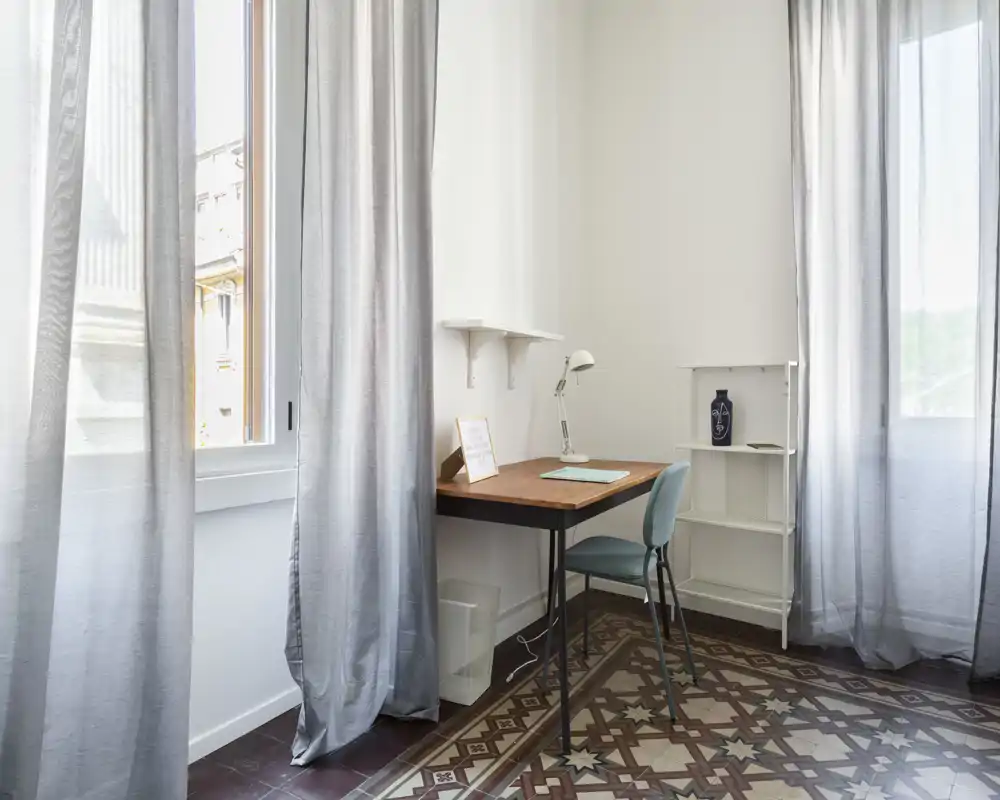 Habitación luminosa y minimalista con escritorio y silla junto a grandes ventanas, ideal como pequeño rincón de estudio u oficina en casa.