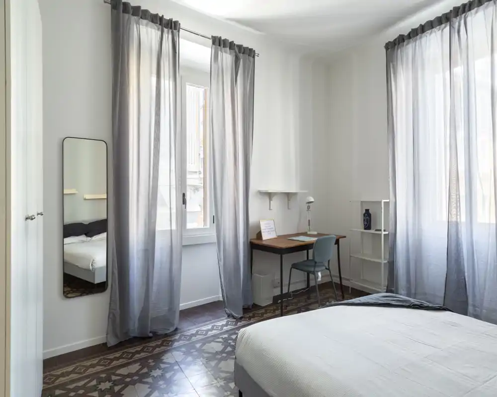 Dormitorio luminoso y aireado con cama grande, suelo de baldosas decoradas y un pequeño escritorio junto a la ventana—ideal para una estancia cómoda y trabajar con luz natural.