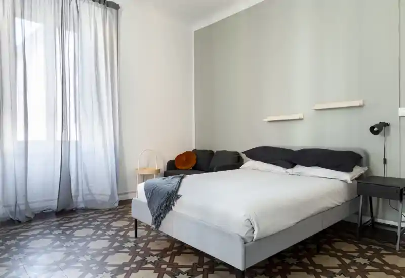 Dormitorio luminoso y con estilo con cama doble bien arreglada, ropa de cama suave y un pequeño asiento junto a la pared; suelo hidráulico decorado y gran ventana con cortinas translúcidas que aportan una sensación aireada.