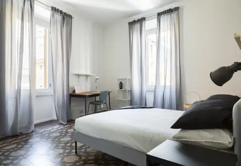 Dormitorio luminoso y despejado con grandes ventanas, cama doble bien hecha y un escritorio pequeño — ideal para descansar y trabajar.