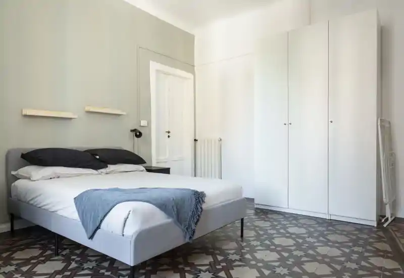 Dormitorio luminoso y minimalista con una cama doble bien hecha, cabecero gris y un gran armario blanco. El suelo de baldosas decoradas y los detalles sencillos aportan carácter y una sensación contemporánea y tranquila.