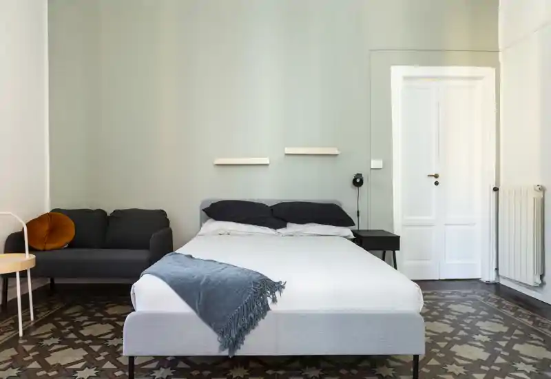 Dormitorio luminoso y minimalista con cama doble centrada, tonos grises suaves, suelo de baldosas decorativas y un pequeño sofá al lado: un espacio de descanso tranquilo y bien iluminado.