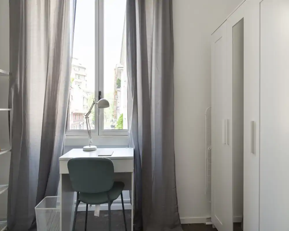 Coin chambre lumineux et minimaliste avec bureau près de la fenêtre, rideaux gris et armoire blanche — parfait pour un studio ou une personne seule.