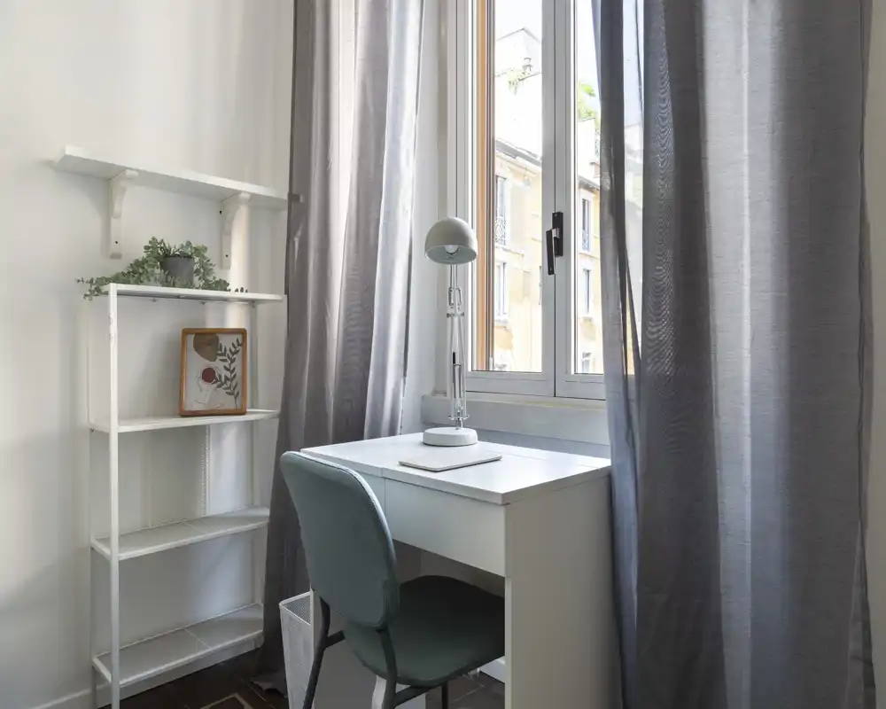 Un petit espace de travail lumineux près de la fenêtre avec un bureau blanc simple, une chaise verte rembourrée et une lampe de bureau ; des voilages filtrent la lumière naturelle et une étagère fine apporte du rangement décoratif.