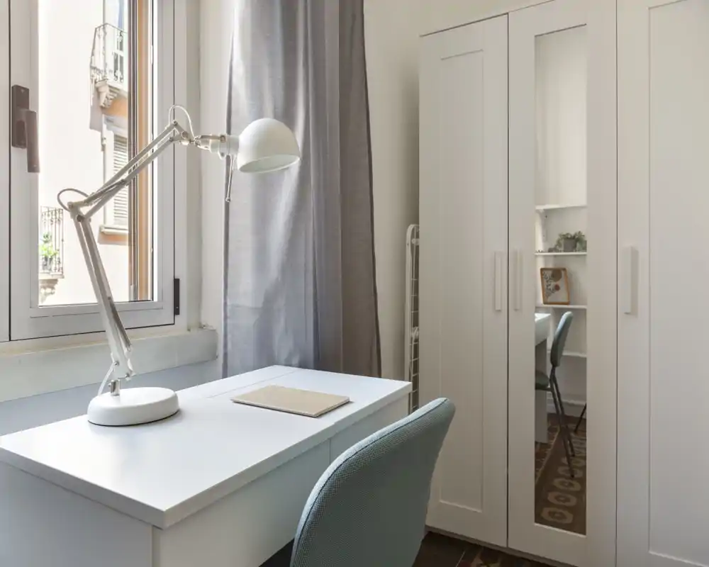Coin de chambre compact avec bureau blanc, lampe de lecture et armoire à porte miroir ; lumière naturelle vive et chaise rembourrée créent un espace de travail tranquille dans la chambre.