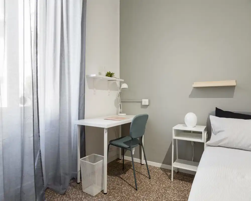Rincón de dormitorio luminoso y minimalista con cama individual, mesita y un pequeño escritorio junto a la ventana. Estilo moderno y luz natural suave que crean un área de descanso y trabajo tranquila.