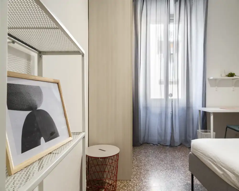 Rincón de dormitorio luminoso y sencillo con cama visible, cortinas suaves y estantería ordenada — decoración minimal ideal para una persona o estancias cortas.