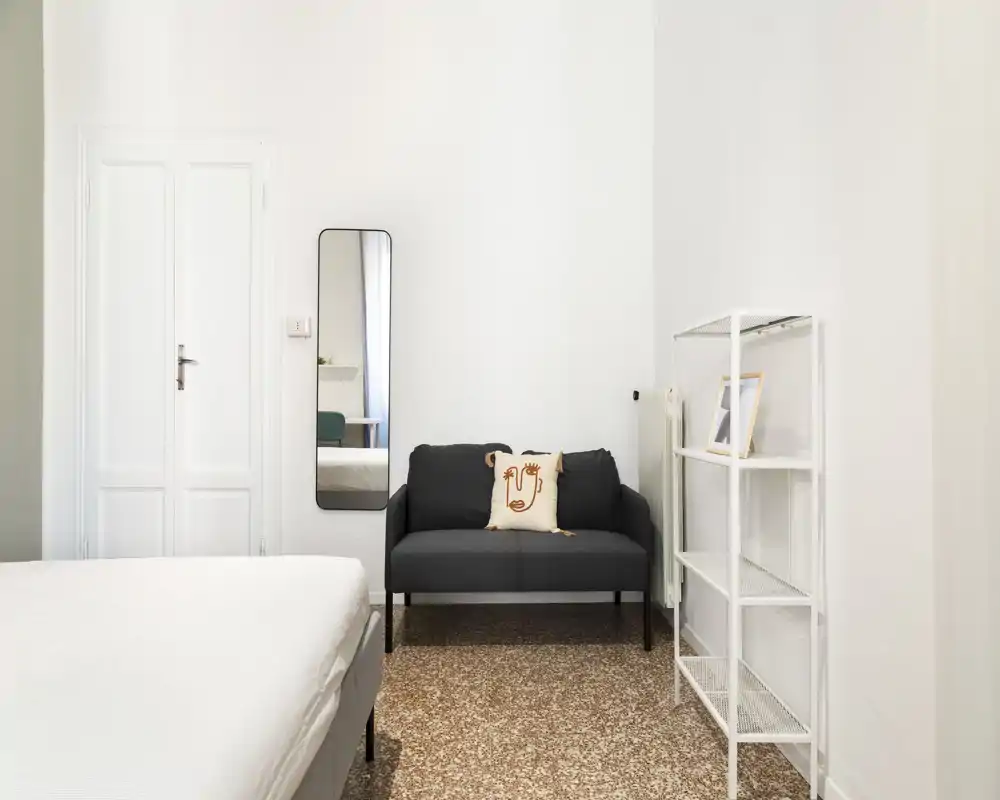 Dormitorio luminoso y minimalista con la cama visible en primer plano, un pequeño sofá gris oscuro, espejo de cuerpo entero y estanterías abiertas; líneas limpias y decoración neutra para un ambiente sereno.