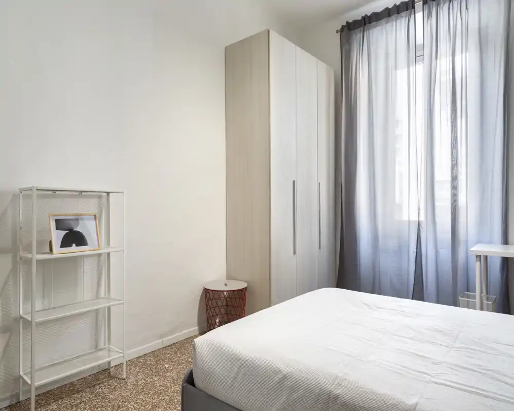 Dormitorio minimalista y luminoso con cama doble bien hecha, armario alto, estantería simple y ventana con cortina translúcida que deja pasar la luz natural.