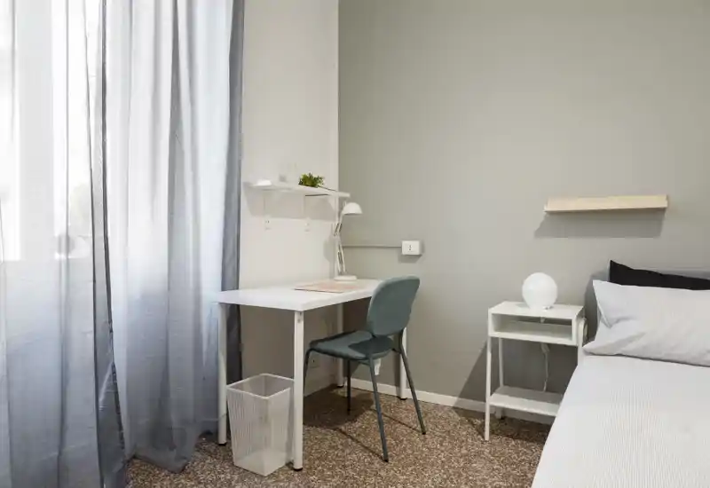 Rincón de dormitorio luminoso y minimalista con cama individual, mesita y un pequeño escritorio junto a la ventana. Estilo moderno y luz natural suave que crean un área de descanso y trabajo tranquila.