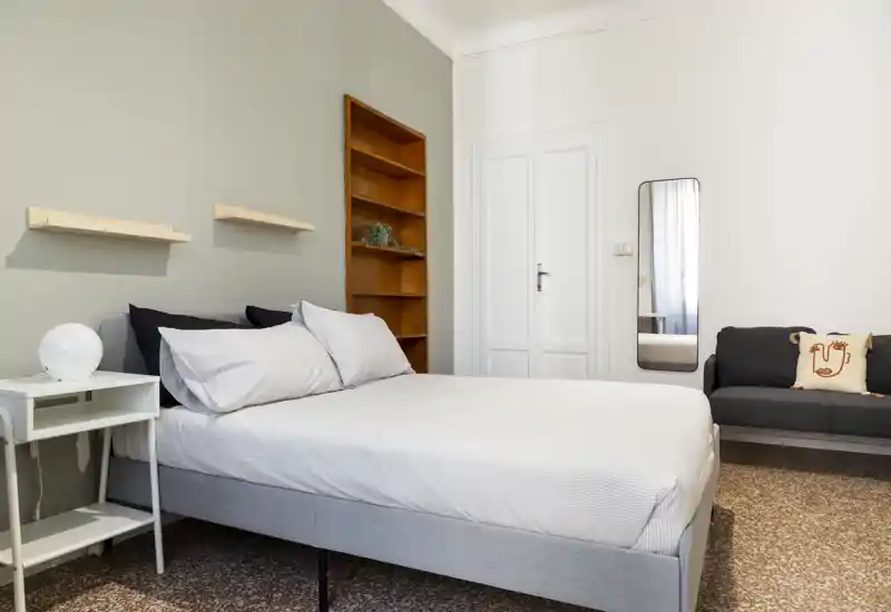 Dormitorio moderno y luminoso con cama doble bien hecha, mesita con lámpara, estantes en la pared y un pequeño sofá — tonos neutros y diseño limpio que lo hacen acogedor.