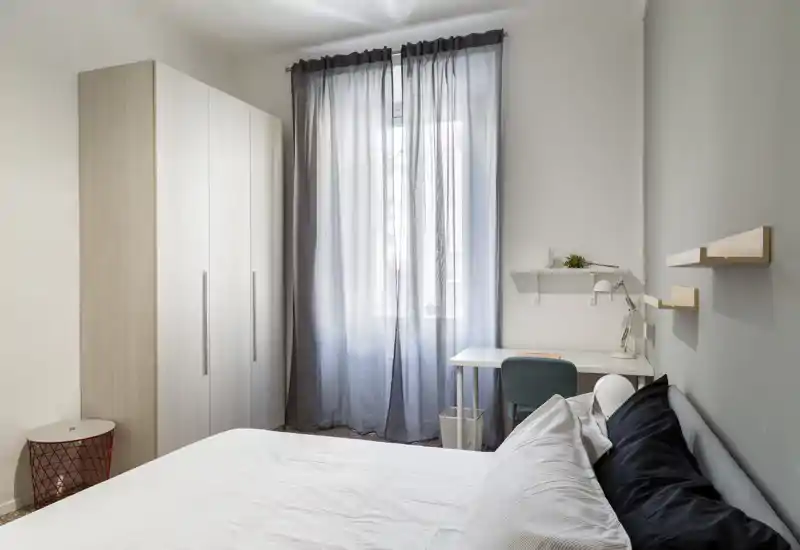 Dormitorio luminoso y ordenado con cama doble, armario y un pequeño escritorio junto a la ventana — tonos neutros y textiles suaves que generan un ambiente tranquilo.