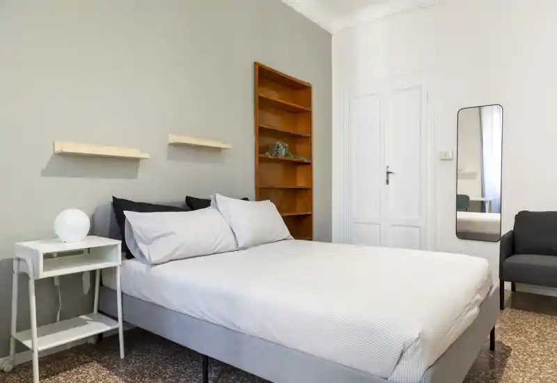 Dormitorio luminoso y bien presentado con cama doble, mesita y estanterías sencillas; tonos neutros y un espejo alto crean un ambiente tranquilo y aireado, perfecto para un anuncio de alquiler.