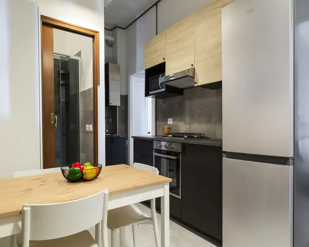 Cocina compacta y moderna con muebles bajos oscuros, armarios altos de madera clara, horno empotrado y frigorífico de acero inoxidable. Una pequeña mesa de madera con sillas y un cuenco de frutas crea un rincón comedor acogedor y funcional.