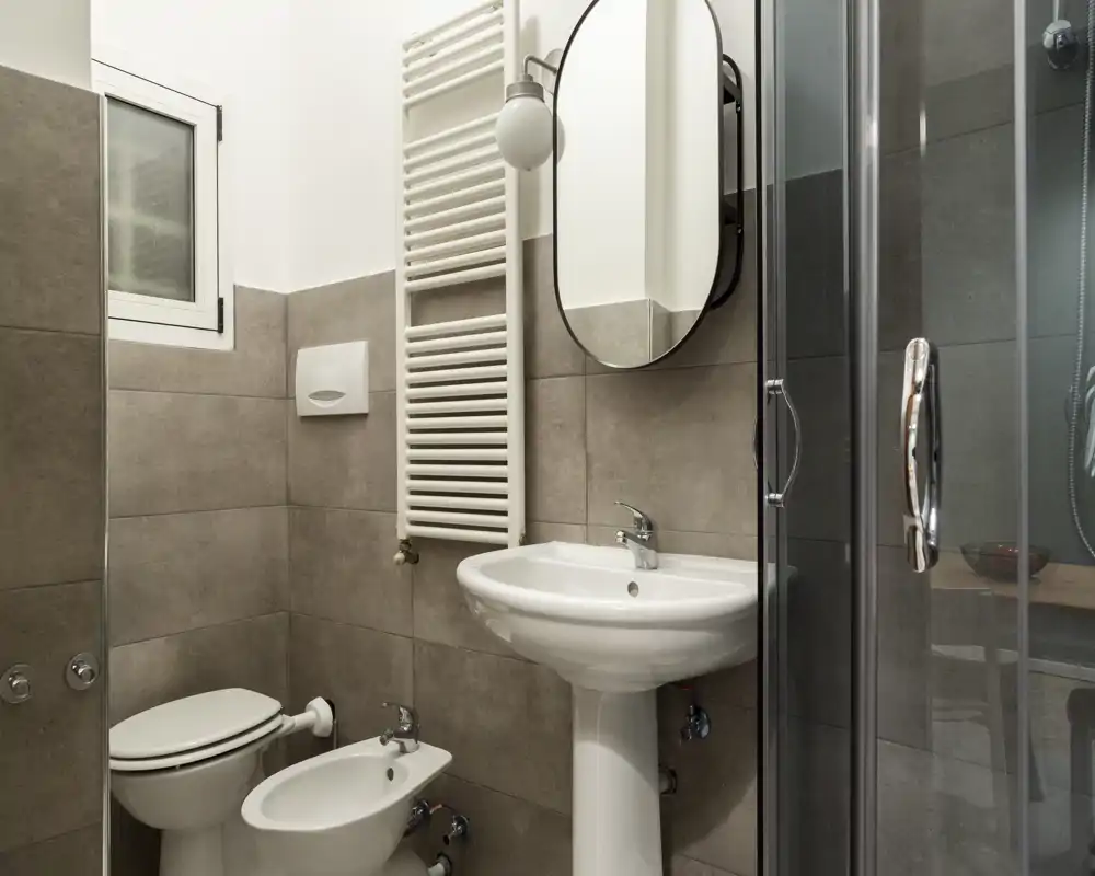 Baño moderno y limpio con lavabo pedestal, bidé, inodoro y mampara de ducha de vidrio. Baldosas grises neutras y radiador toallero mural que aportan un estilo contemporáneo.
