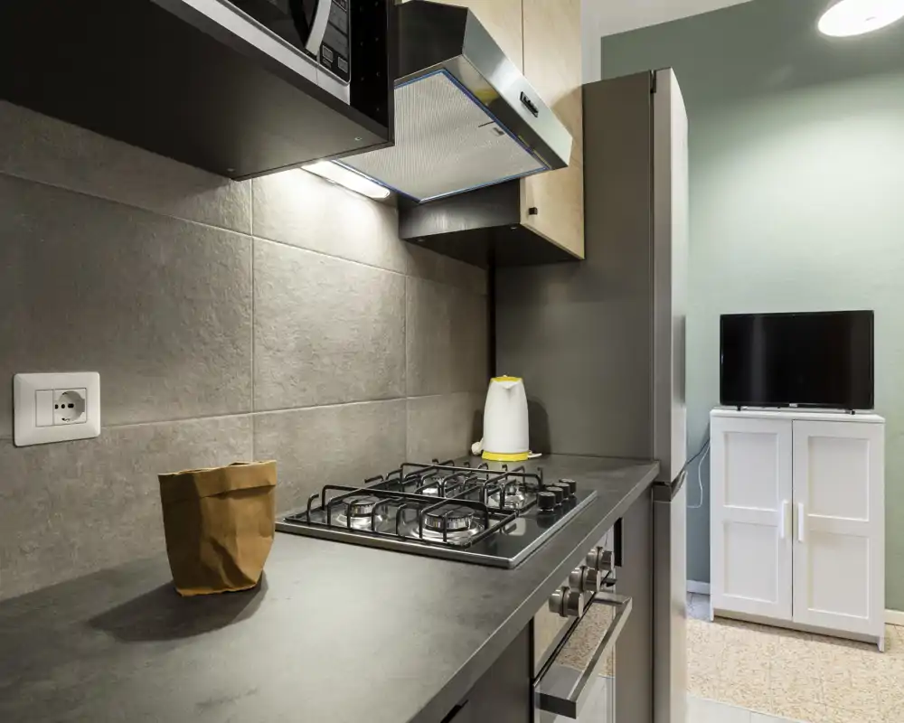 Cocina moderna y compacta con placa de gas, luz bajo armarios y electrodomésticos de acero; encimera ordenada con hervidor y decoración mínima.