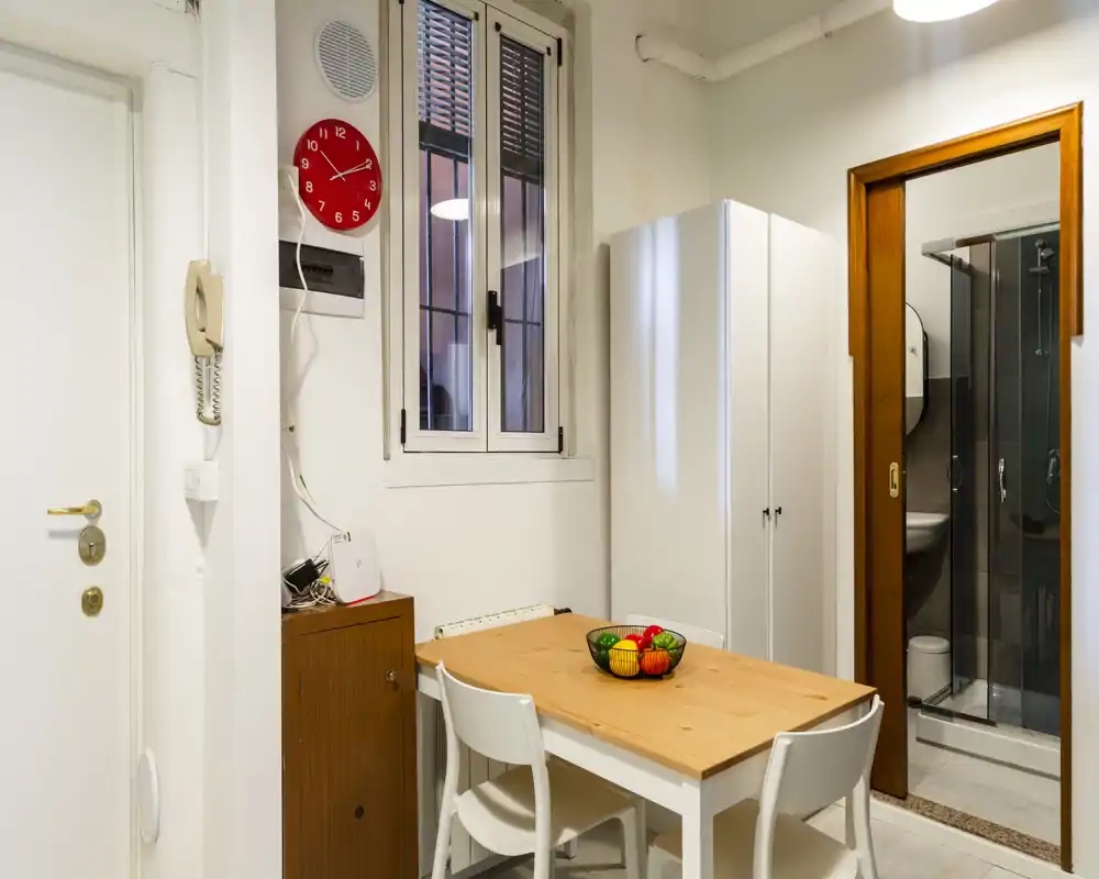 Rincón comedor compacto con mesa de madera, cuatro sillas y un cuenco de frutas; paredes neutras y un armario blanco crean un área de comedor práctica y ordenada junto al baño.