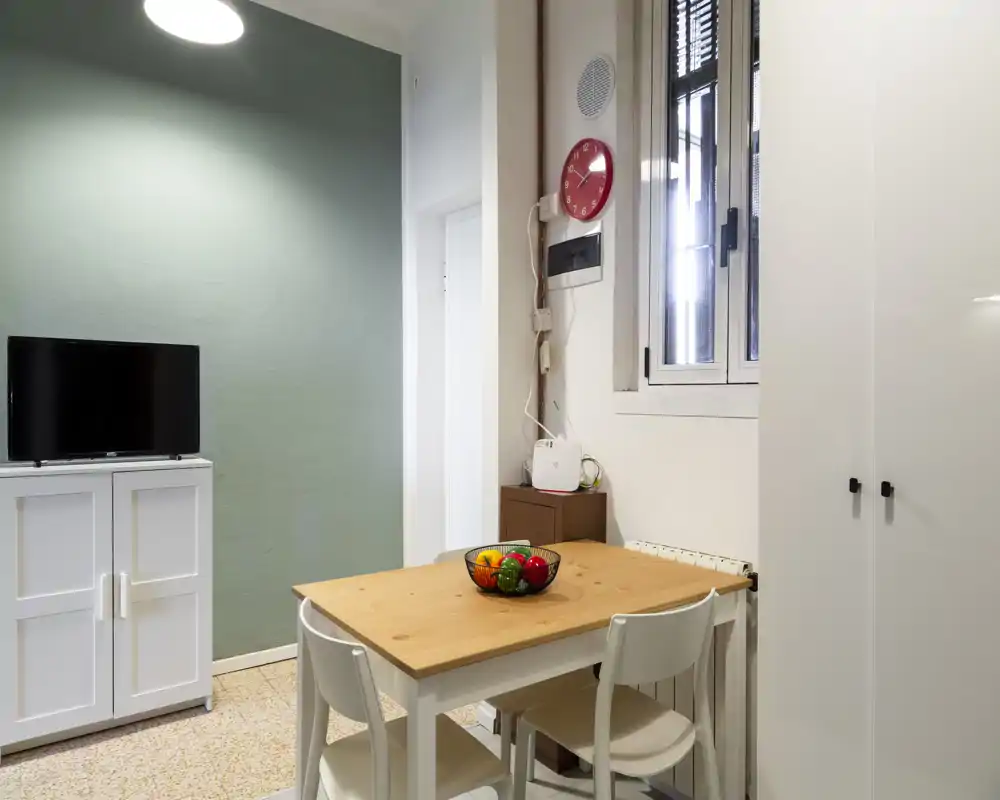 Zona de comedor compacta con mesa de madera, cuatro sillas y un cuenco de frutas decorativo; espacio luminoso y ordenado con mueble TV y armario visibles, perfecto para apartamentos pequeños.