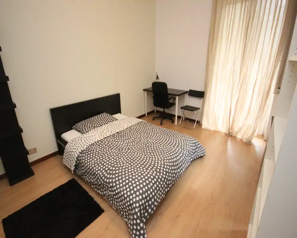 Camera da letto luminosa e minimal con un letto matrimoniale a fantasia, scrivania e scaffalatura; pavimento in legno chiaro e luce naturale rendono l'ambiente accogliente.