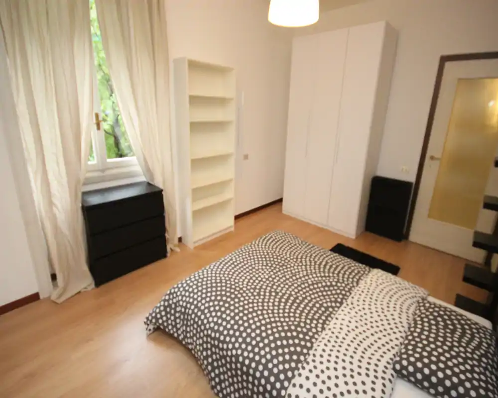 Camera da letto luminosa e arredata semplicemente con un letto matrimoniale con copriletto a fantasia, un armadio bianco, mensole e luce naturale dalla finestra con tende.