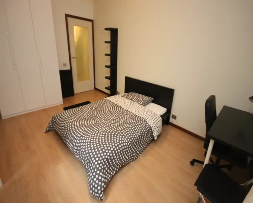 Camera da letto luminosa e arredata in modo semplice con letto matrimoniale, armadio e scrivania; pavimento in legno e colori neutri per un aspetto pulito e moderno.