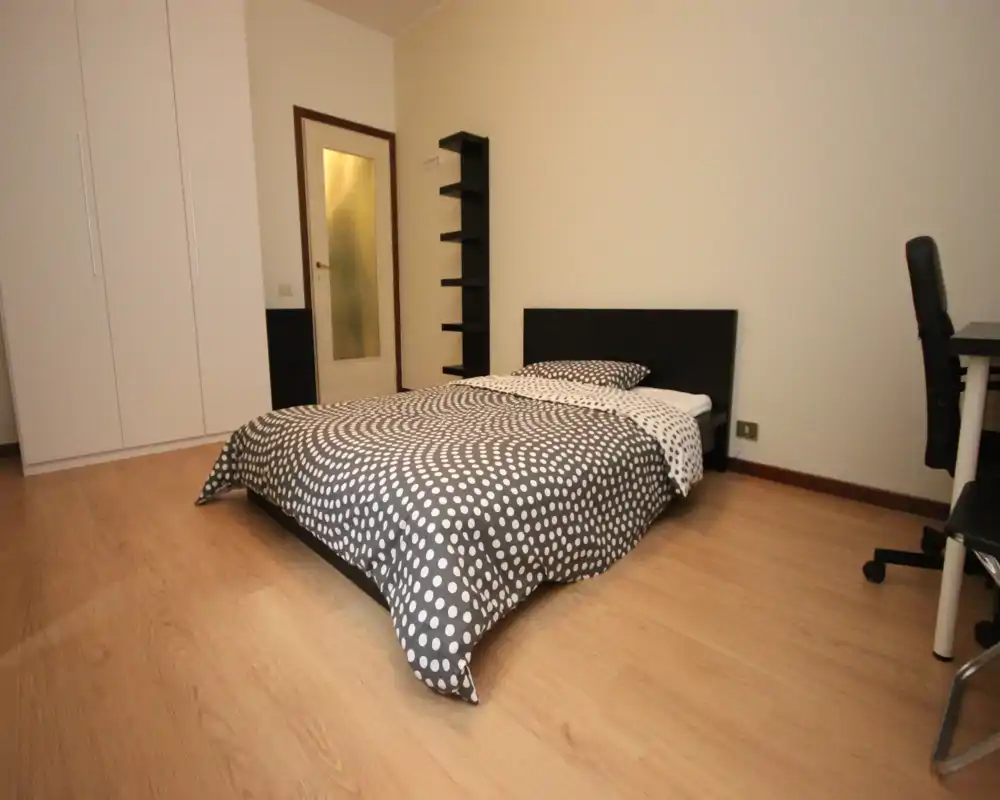 Camera da letto luminosa e arredata in modo semplice con letto matrimoniale con copriletto a pois, armadio, mensole e una piccola scrivania — ideale per un soggiorno confortevole.