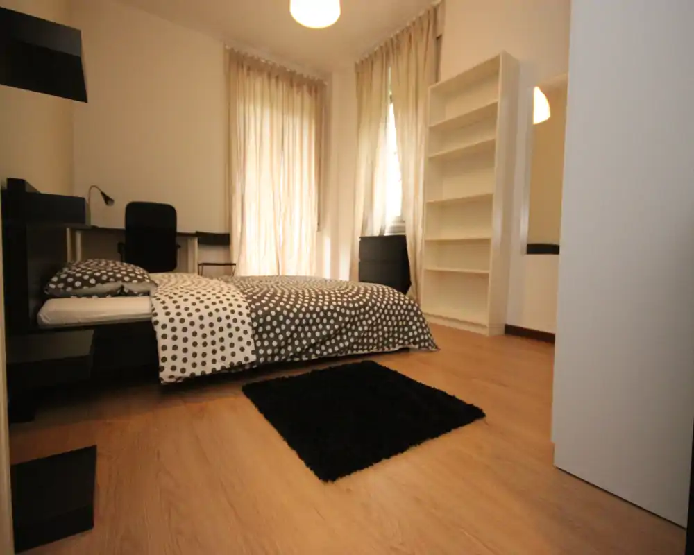 Camera da letto moderna e luminosa con copriletto a pois, pavimento in legno e piccolo tappeto nero; grandi finestre con tende chiare e un angolo scrivania sullo sfondo.