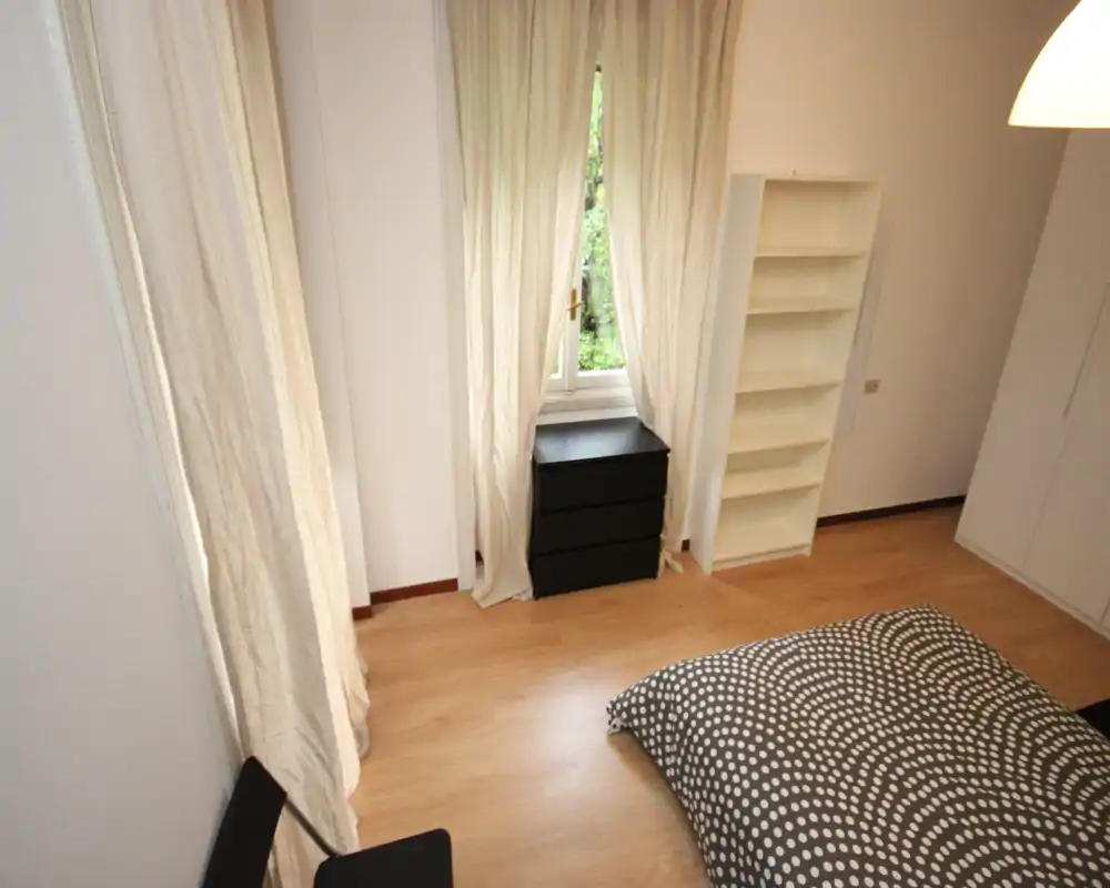 Camera da letto luminosa e arredata in modo semplice con una coperta a fantasia, cassettiera sotto la finestra e una libreria a giorno. La luce naturale e le tende chiare creano un'atmosfera neutra e rilassante.