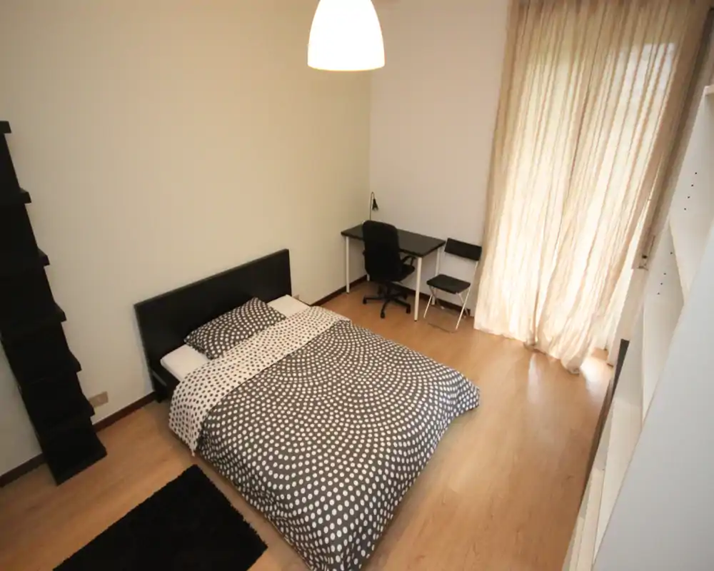 Camera da letto moderna e luminosa con ampio letto matrimoniale, copriletto a fantasia, scrivania e luce naturale soffusa dalle tende a tutta altezza.