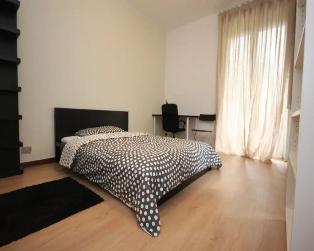 Camera da letto luminosa e minimal con letto matrimoniale, copriletto a pois, scrivania e luce naturale da tende a pacchetto lunghe fino al pavimento.