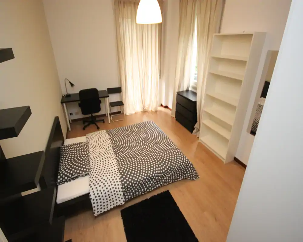 Camera da letto moderna e luminosa con letto matrimoniale a fantasia, pavimento in legno, scrivania e scaffalature; la luce naturale filtrata dalle tende crea un'atmosfera tranquilla.