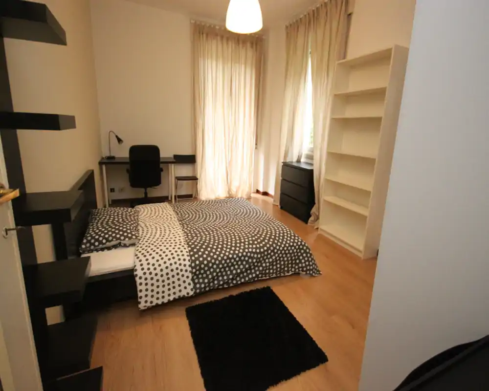 Camera da letto luminosa e moderna con un letto matrimoniale con biancheria a fantasia, una scrivania vicino alla finestra, scaffalature e pavimento in legno chiaro—ben arredata e ariosa.