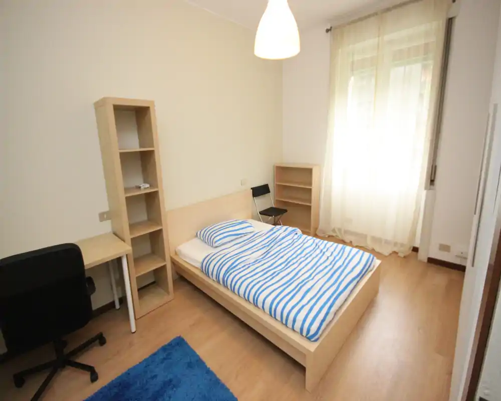 Bright, simply furnished bedroom with a double bed, desk and shelving; natural light from a large window and wooden flooring create a clean, airy feel.