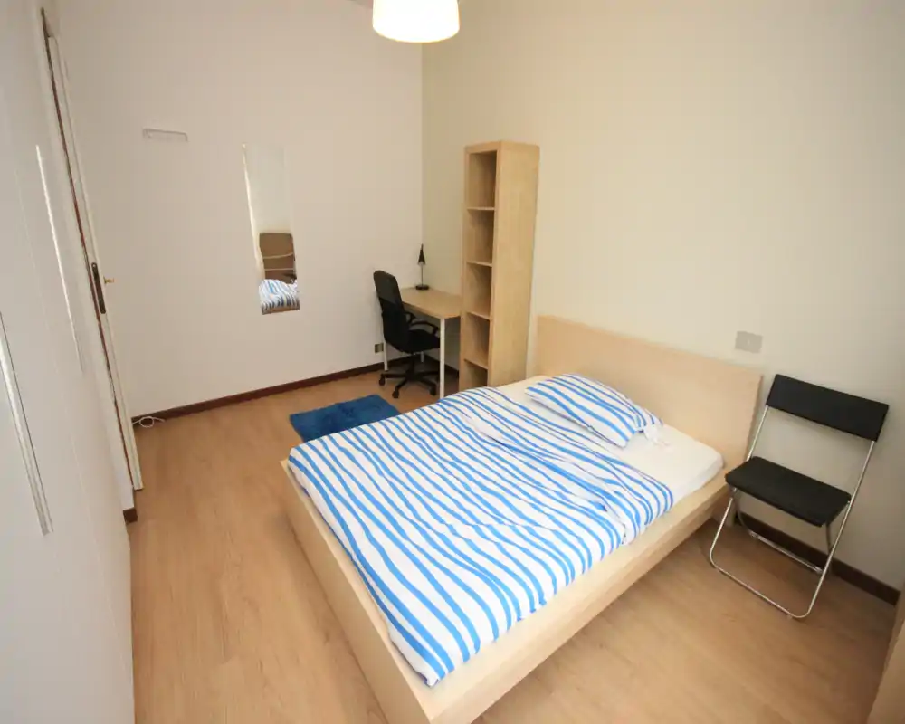 Bright, tidy bedroom with a double bed dressed in blue-striped linens, a small desk and shelving unit — ideal for a single occupant or couple. Clean wooden floor and minimal decor give a calm, functional feel.
