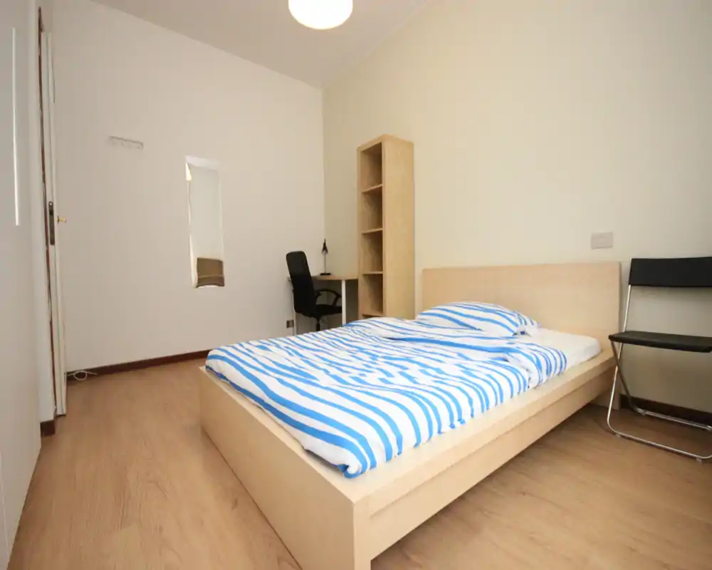 Minimal, well-lit bedroom with a light wood platform bed, striped bedding and simple furniture including a desk and shelving — clean and functional space ideal for a single occupant.