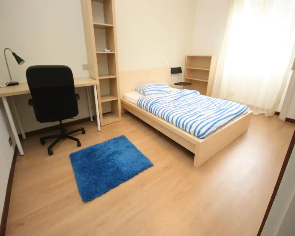 Bright, minimalist single bedroom with a wooden bed frame, striped bedding, desk and chair; natural light from a large window and a small blue rug add warmth.