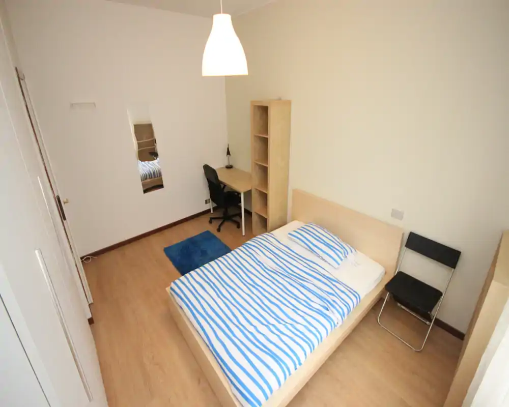 Bright, minimalist bedroom with a double bed dressed in blue-and-white striped linens, a small desk and shelving unit, and a simple chair — ideal for a single occupant or couple seeking a clean, functional space.
