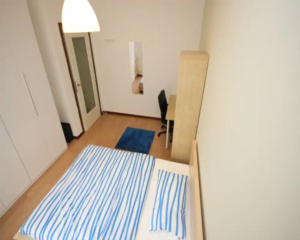 Bright, simply furnished bedroom photographed from a high angle showing a double bed with striped bedding, wardrobe, desk and a small blue rug.