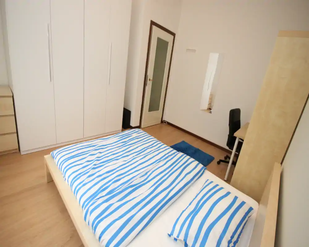 Bright, minimal bedroom with a double bed dressed in blue-and-white striped bedding, a large white wardrobe, desk and simple wooden flooring—clean and inviting for short-term stays.