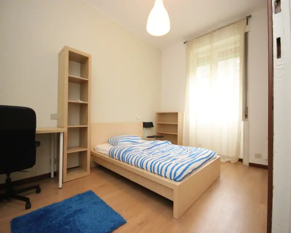Simple, bright single bedroom with a wooden platform bed, striped bedding, natural light from a large window and a small desk and shelving — clean and modern minimal setup ideal for a student or lone occupant.