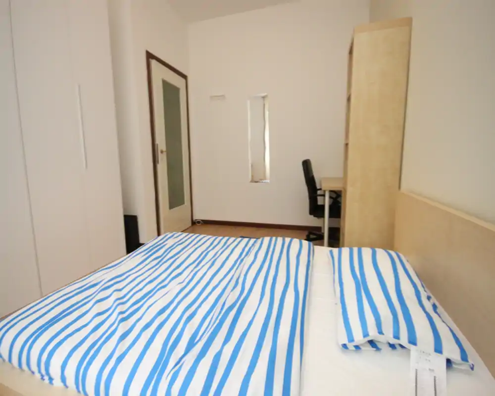 Bright, simply furnished bedroom with a double bed dressed in blue-and-white striped linens, a wardrobe and a small desk with chair. Neutral walls and minimal decor create a clean, airy atmosphere ideal for short-term rentals.